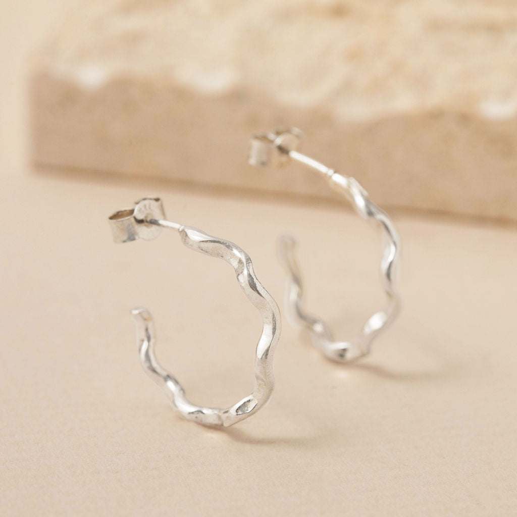 Sterling silver wavy hoop earrings with an organic molten texture, handmade by Rockpool Jewellery and styled on a neutral stone background