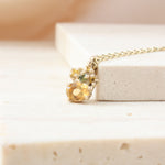 A delicate gold chain necklace with a multi-stone pendant resting on a light beige stone block. The pendant features a large round yellow gemstone, a smaller round yellow gemstone, and a small green gemstone, all set in a gold granules