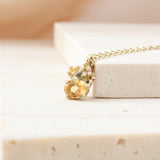 A delicate gold chain necklace with a multi-stone pendant resting on a light beige stone block. The pendant features a large round yellow gemstone, a smaller round yellow gemstone, and a small green gemstone, all set in a gold granules