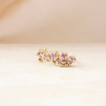 A gold-toned textured ring with a row of small pink gemstones, resting on a light beige surface with a soft beige background.