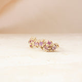 A gold-toned textured ring with a row of small pink gemstones, resting on a light beige surface with a soft beige background.
