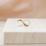 A pair of minimalist gold and silver hoop earrings with small gold rockpool design, displayed on a light beige stone block against a soft beige background.