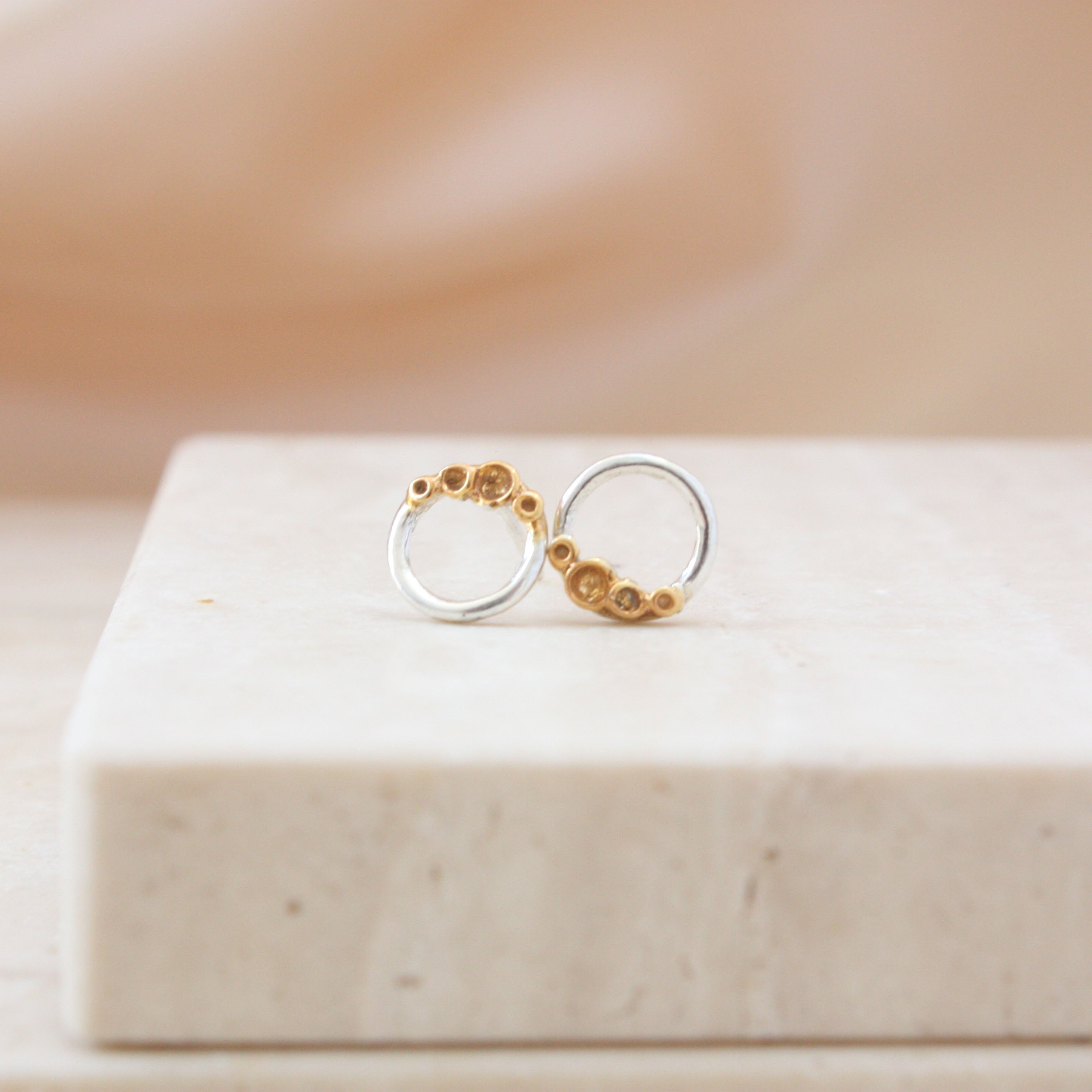 A pair of minimalist gold and silver hoop earrings with small gold rockpool design, displayed on a light beige stone block against a soft beige background.