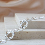 A close-up of a silver chain bracelet with textured, wavy open-link rings, draped over stacked beige stone blocks against a soft beige fabric background.