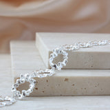 A close-up of a silver chain bracelet with textured, wavy open-link rings, draped over stacked beige stone blocks against a soft beige fabric background.