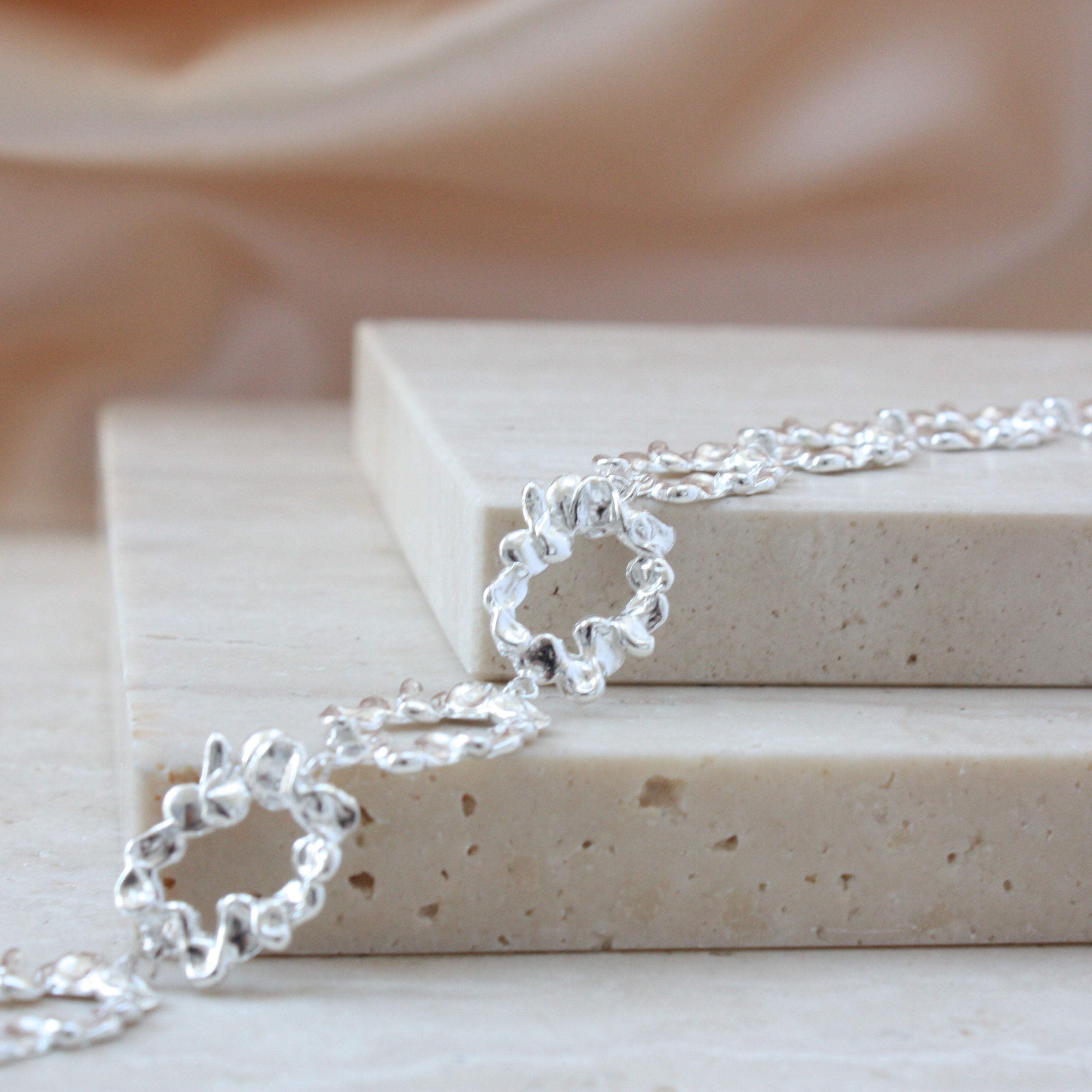 A close-up of a silver chain bracelet with textured, wavy open-link rings, draped over stacked beige stone blocks against a soft beige fabric background.