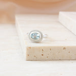 A silver ring with an oval light blue gemstone, set in a textured bezel, resting on a beige stone block.