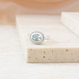 A silver ring with an oval light blue gemstone, set in a textured bezel, resting on a beige stone block.