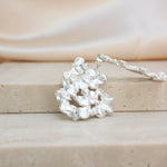 A silver-toned textured earring with a cluster of irregular leaf-like or coral-like elements displayed on a beige stone block with a soft cream fabric background.