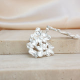 A silver-toned textured earring with a cluster of irregular leaf-like or coral-like elements displayed on a beige stone block with a soft cream fabric background.