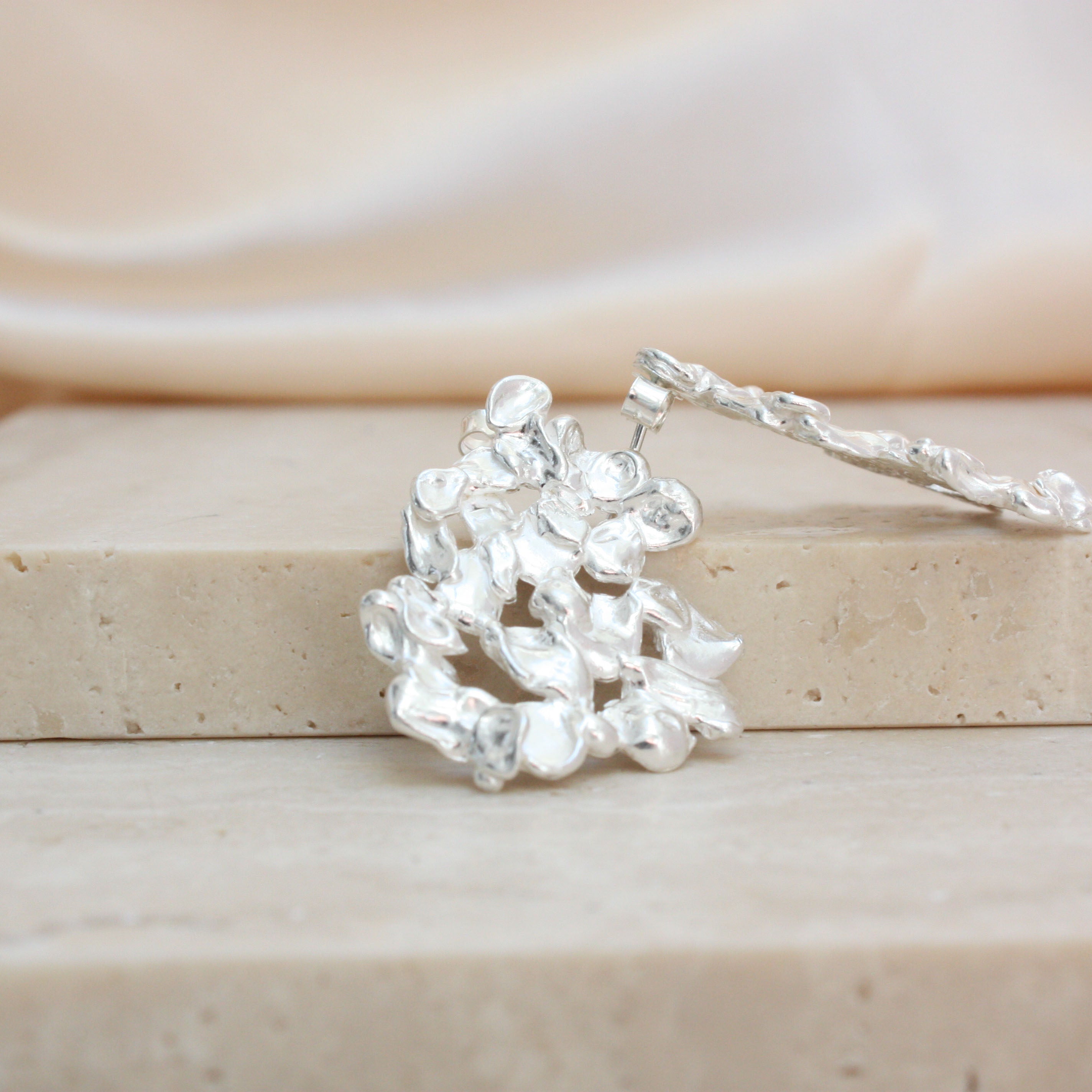 A silver-toned textured earring with a cluster of irregular leaf-like or coral-like elements displayed on a beige stone block with a soft cream fabric background.