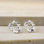 A pair of small silver hoop earrings with a textured, wavy, organic outline, displayed on a light beige stone surface.