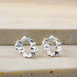A pair of small silver hoop earrings with a textured, wavy, organic outline, displayed on a light beige stone surface.