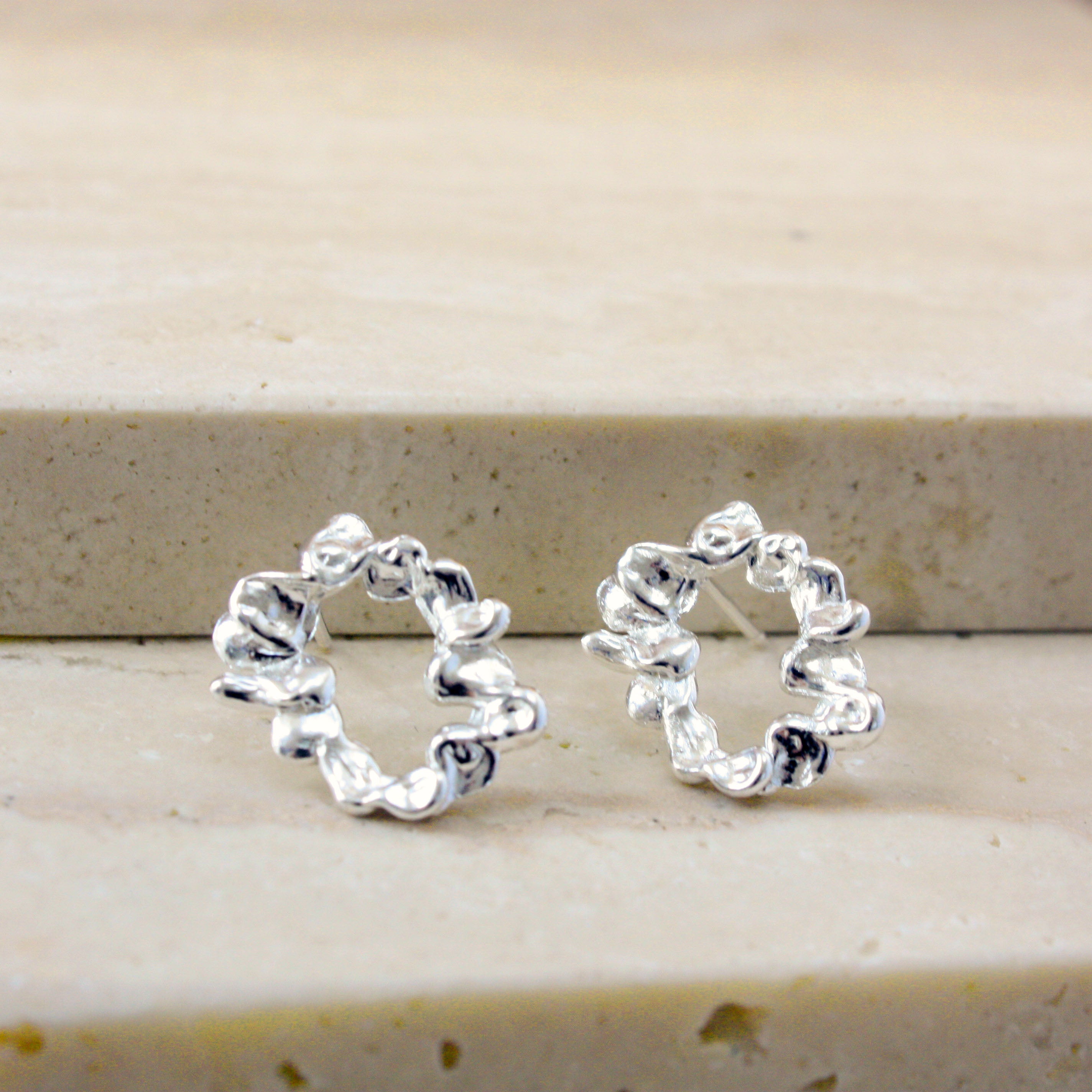 A pair of small silver hoop earrings with a textured, wavy, organic outline, displayed on a light beige stone surface.